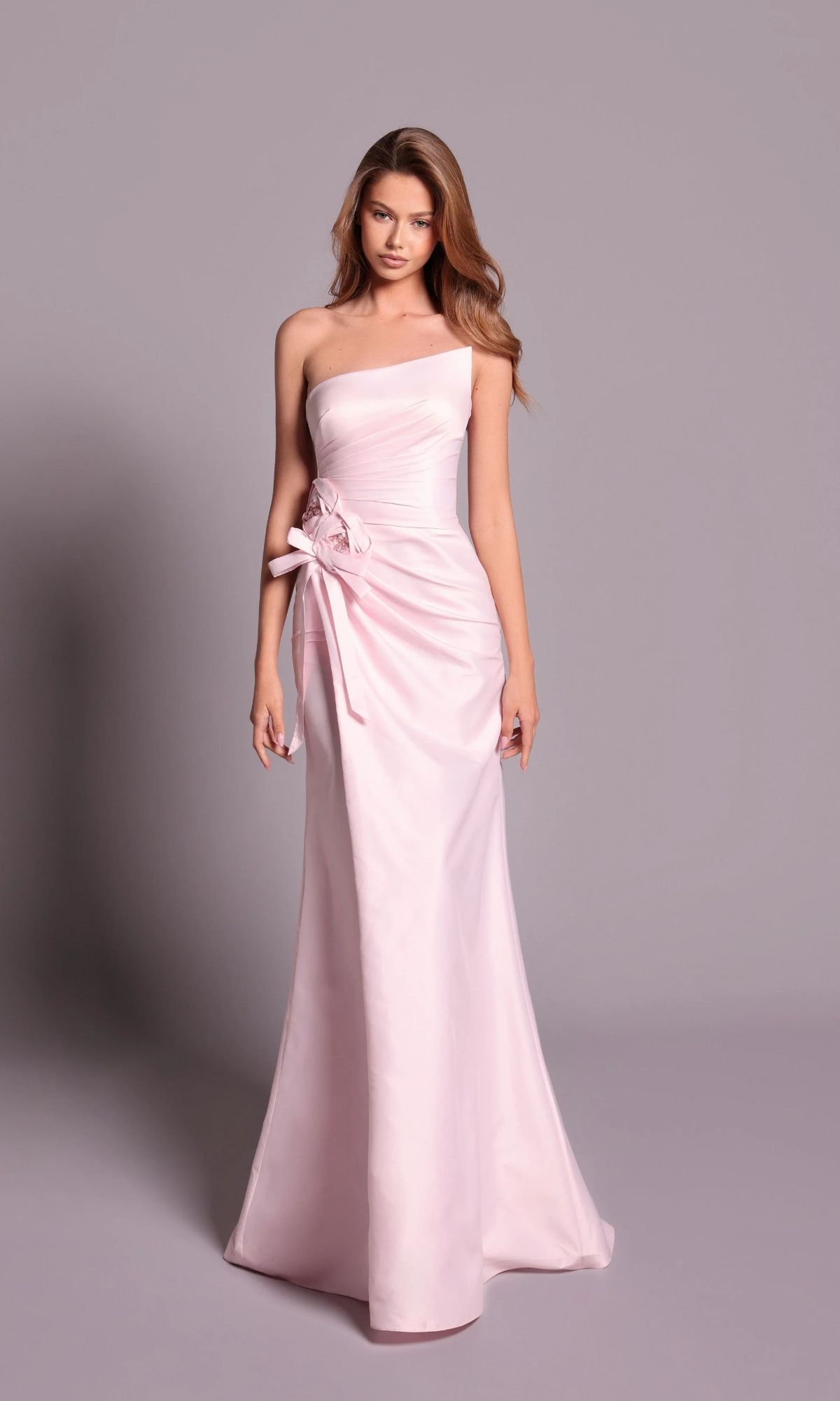 A woman stands in front of a plain grey background wearing the Tarik Ediz 99126— a light pink, strapless, floor-length formal dress featuring a floral detail at the waist.